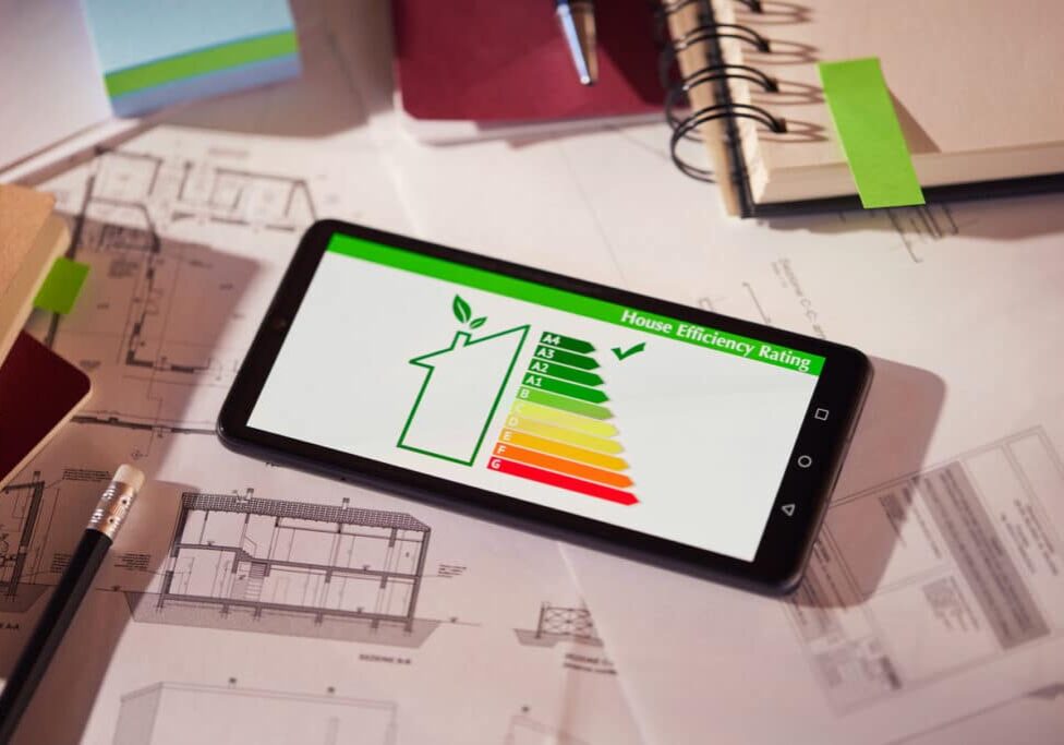 Energy efficiency mobile app on screen. Closeup of detail of house efficiency rating on mobile phone. Home efficiency reading app on mobile phone kept on desk. Energy efficiency screen on cellphone leaning on drawings and blueprints of a new house. Architect's desk with plans on it and a cell phone with an energy efficiency graph. Renewable energy, renovation, energy class and eco house concept.
