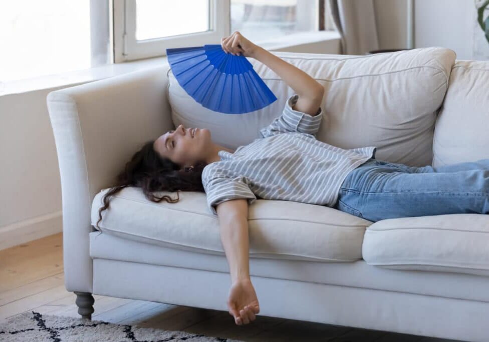 Exhausted young woman suffering from hot weather, overheat Exhausted young woman suffering from hot weather, overheat, too high air temperature at home, lying on sofa, waving paper handheld fan, cooling, feeling discomfort